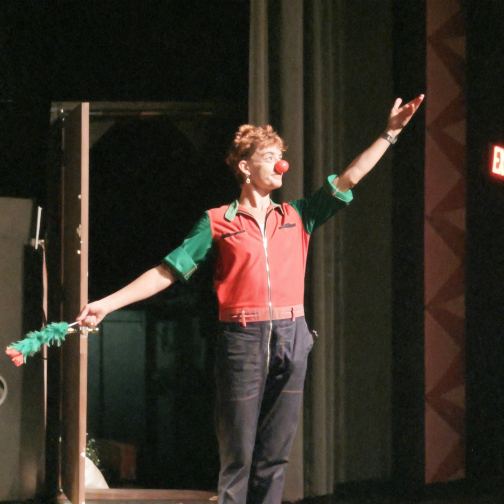 Clown Daddy, also known as Ellie J, performing on stage with a red clown nose and a green feather duster